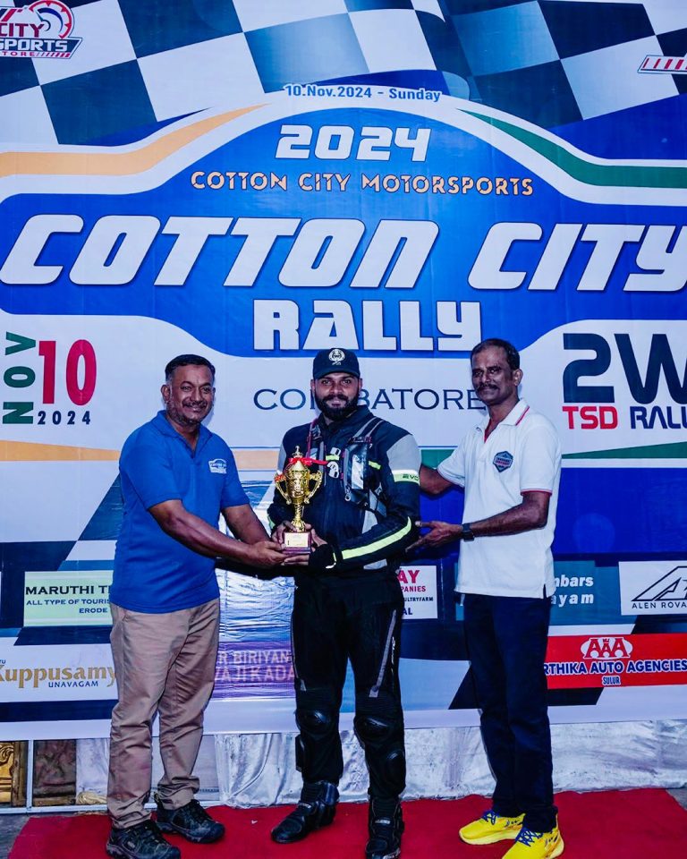Three men celebrate at the Cotton City Rally, with one in racing gear holding a trophy, all against a vibrant event backdrop.